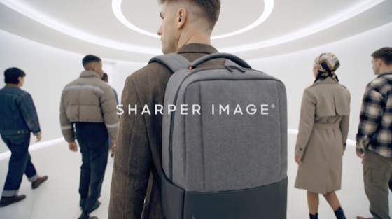 Fotograma de un video de producto: personas caminando por un pasillo blanco, con una mochila gris en primer plano