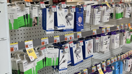 Pared de una tienda con decenas de cables y cargadores de varias marcas colgados en ganchos, con etiquetas de precio y de oferta