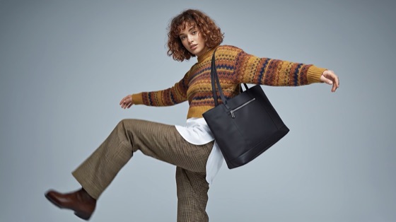 Modelo con un suéter tejido de colores y una cartera tote negra al hombro, con una pierna levantada sobre un fondo gris azulado